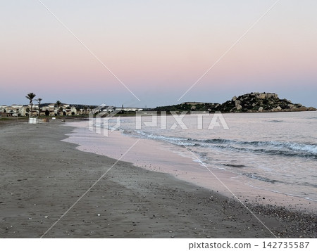 Quiet sandy beach with gentle waves, rocky shoreline, coastal houses, and pastel evening sky Quiet sandy beach with gentle waves, rocky shoreline, coastal houses, and pastel evening sky 142735587