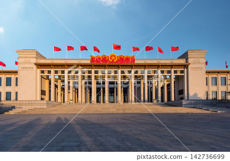 Grand symmetrical government building in Tiananmen Square, Beijing, with red flags and golden banner under blue sky. Grand symmetrical government building in Tiananmen Square, Beijing, with red flags and golden banner under blue sky. 142736699