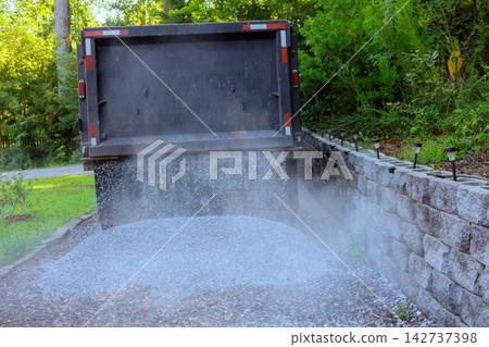 Dump truck unloads gravel in residential yard near stone wall 142737398