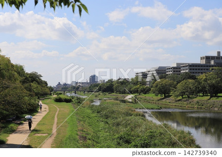 京都鴨川河畔的夏日景色,藍天白雲,綠樹成蔭的步道,視野開闊。可作為旅遊宣傳和健康步行廣告的背景資料。 京都鴨川河畔的夏日景色,藍天白雲,綠樹成蔭的步道,視野開闊。可作為旅遊宣傳和健康步行廣告的背景資料。 142739340