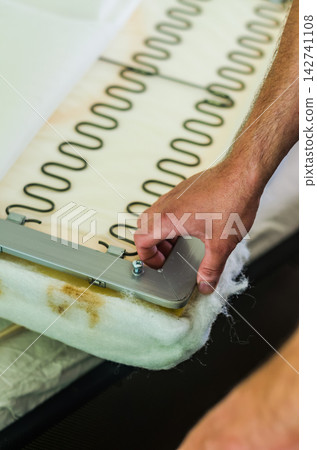 Upholsterer repairing furniture. Close-up of male hands installing zig-zag sinuous springs on a sofa frame with foam padding and white batting. . High quality photo Upholsterer repairing furniture. Close-up of male hands installing zig-zag sinuous springs on a sofa frame with foam padding and white batting. . High quality photo 142741108