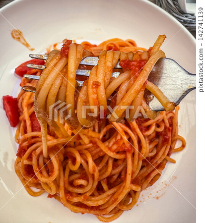 Close-up of fork twirling spaghetti pasta coated in rich tomato sauce, served on white plate with fresh tomato slices, authentic Italian cuisine. High quality photo Close-up of fork twirling spaghetti pasta coated in rich tomato sauce, served on white plate with fresh tomato slices, authentic Italian cuisine. High quality photo 142741139