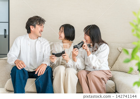 一家三口,笑容滿面,玩電子遊戲玩得不亦樂乎。他們正享受著輕鬆愉快的家庭假期。 一家三口,笑容滿面,玩電子遊戲玩得不亦樂乎。他們正享受著輕鬆愉快的家庭假期。 142741144