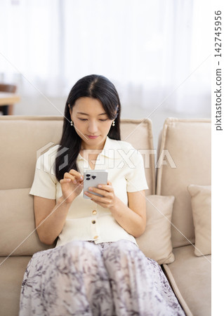 一位年輕的日本女性正在沙發上使用智慧型手機。 142745956