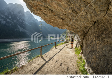 Hikers and dog walk along Fore Gosau Lake scenic path underneath a rock overhang Hikers and dog walk along Fore Gosau Lake scenic path underneath a rock overhang 142746391