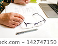 Close-up of a person hands holding glasses on a desk with documents and a laptop. Office work, business, and paperwork concept 142747150