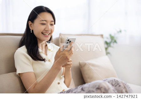一位年輕的日本女子坐在沙發上，一邊使用智慧型手機一邊微笑。 142747921