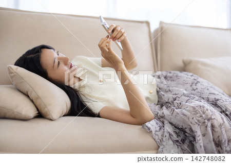 A young Japanese woman is lying on a sofa and using her smartphone. A young Japanese woman is lying on a sofa and using her smartphone. 142748082