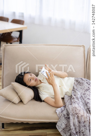 A young Japanese woman is lying on a sofa and using her smartphone. A young Japanese woman is lying on a sofa and using her smartphone. 142748087