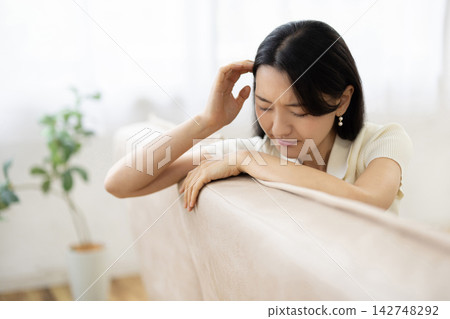 一位年輕的日本女子坐在明亮的窗邊沙發上，摸著頭，做了個鬼臉。 142748292