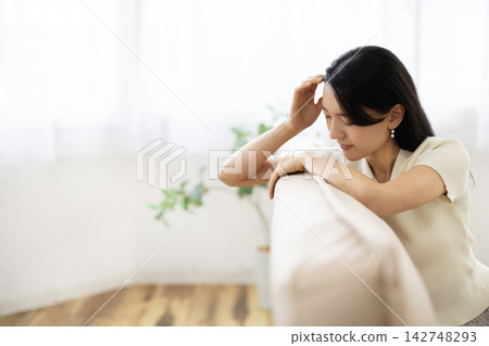 一位年輕的日本女子坐在明亮的窗邊沙發上，摸著頭，做了個鬼臉。 142748293
