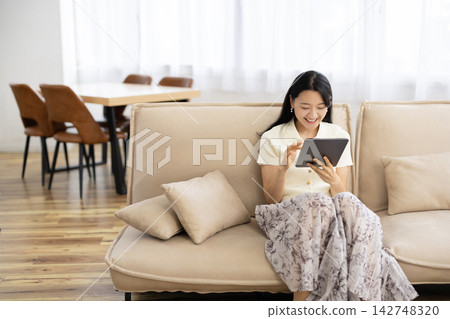 一位年輕的日本女子坐在沙發上，一邊操作平板電腦一邊微笑。 142748320