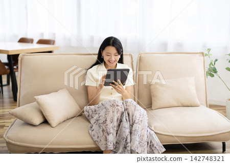A young Japanese woman smiles while operating a tablet while sitting on a sofa. A young Japanese woman smiles while operating a tablet while sitting on a sofa. 142748321