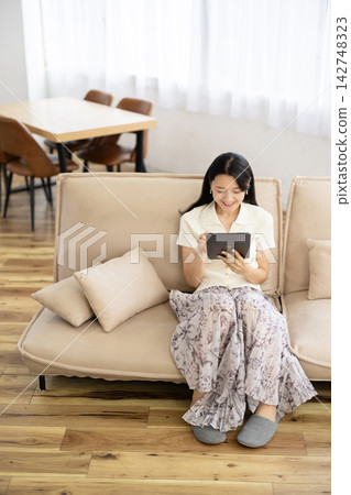 A young Japanese woman smiles while operating a tablet while sitting on a sofa. A young Japanese woman smiles while operating a tablet while sitting on a sofa. 142748323