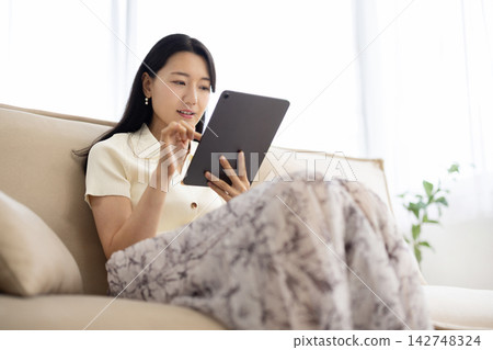 一位年輕的日本女子坐在沙發上，一邊操作平板電腦一邊微笑。 142748324