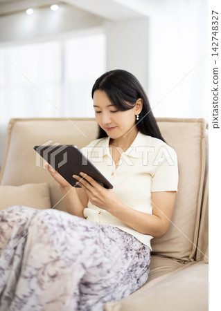 A young Japanese woman smiles while operating a tablet while sitting on a sofa. A young Japanese woman smiles while operating a tablet while sitting on a sofa. 142748327
