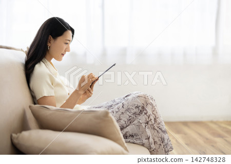 A young Japanese woman smiles while operating a tablet while sitting on a sofa. A young Japanese woman smiles while operating a tablet while sitting on a sofa. 142748328