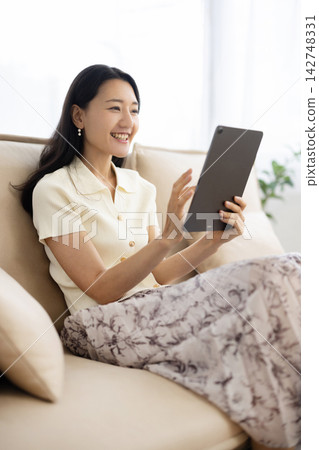 A young Japanese woman smiles while operating a tablet while sitting on a sofa. A young Japanese woman smiles while operating a tablet while sitting on a sofa. 142748331