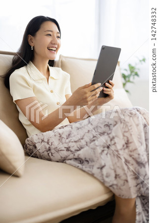 A young Japanese woman smiles while operating a tablet while sitting on a sofa. A young Japanese woman smiles while operating a tablet while sitting on a sofa. 142748332