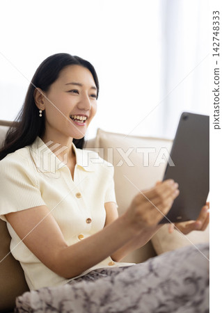 A young Japanese woman smiles while operating a tablet while sitting on a sofa. A young Japanese woman smiles while operating a tablet while sitting on a sofa. 142748333