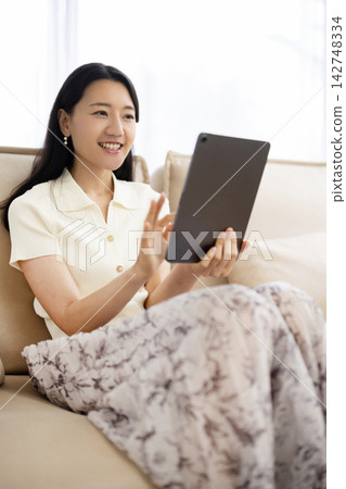 A young Japanese woman smiles while operating a tablet while sitting on a sofa. A young Japanese woman smiles while operating a tablet while sitting on a sofa. 142748334