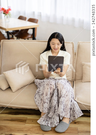 一位年輕的日本女子坐在沙發上，一邊操作平板電腦一邊微笑。 142748335