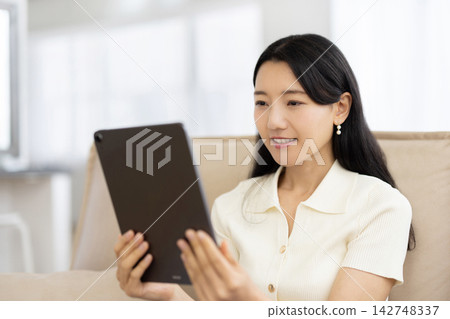 A young Japanese woman smiles while operating a tablet while sitting on a sofa. A young Japanese woman smiles while operating a tablet while sitting on a sofa. 142748337