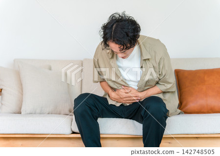 A man is seen clutching his stomach while sitting on a sofa at home. This image suggests he is suffering from a sudden illness such as a stomach ache, stomach pain, or food poisoning. A man is seen clutching his stomach while sitting on a sofa at home. This image suggests he is suffering from a sudden illness such as a stomach ache, stomach pain, or food poisoning. 142748505