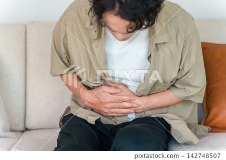 A man is seen on his sofa at home, clutching his stomach in pain. This image suggests he is suffering from severe abdominal pain, stomach ache, or other health problems. A man is seen on his sofa at home, clutching his stomach in pain. This image suggests he is suffering from severe abdominal pain, stomach ache, or other health problems. 142748507