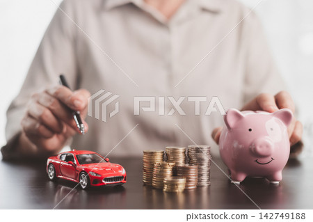 Red model car, pink piggy bank, and coin stacks beside a person with a pen symbolize vehicle savings, purchase budgeting, auto financing, ownership planning, and future financial goals. Red model car, pink piggy bank, and coin stacks beside a person with a pen symbolize vehicle savings, purchase budgeting, auto financing, ownership planning, and future financial goals. 142749188