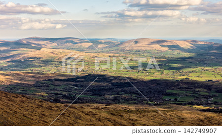 A panoramic view shows rolling hills and mountains under a partly cloudy sky, with some areas illuminated by sunlight. A panoramic view shows rolling hills and mountains under a partly cloudy sky, with some areas illuminated by sunlight. 142749907