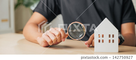 A man hand holds a magnifying glass inspecting a cottage house. Represents real estate appraisal, home inspection, and property assessment for search and investment A man hand holds a magnifying glass inspecting a cottage house. Represents real estate appraisal, home inspection, and property assessment for search and investment 142750927