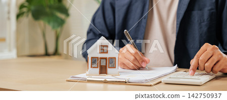 A man writes a signature on house insurance documents, using a calculator for tax and property details. Counting payments and rent by model house, he finalizes the price A man writes a signature on house insurance documents, using a calculator for tax and property details. Counting payments and rent by model house, he finalizes the price 142750937