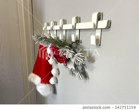 A red Santa hat with white trim hangs on a row of white hooks. A red Santa hat with white trim hangs on a row of white hooks. 142751159