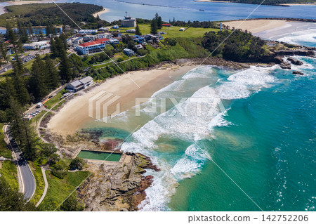 Aerial view of Yamba Main Beach and the ocean pool, with Yamba Lighthouse, the coastal headland and Clarence River visible in the background in New South Wales, Australia. Aerial view of Yamba Main Beach and the ocean pool, with Yamba Lighthouse, the coastal headland and Clarence River visible in the background in New South Wales, Australia. 142752206