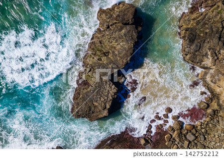 Downward aerial view of ocean waves crashing onto rugged rock formations along the Yamba coastline, creating white foam and natural patterns beside the Pacific Ocean. Downward aerial view of ocean waves crashing onto rugged rock formations along the Yamba coastline, creating white foam and natural patterns beside the Pacific Ocean. 142752212