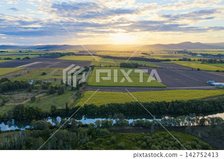 Aerial view of sunset over sugar cane fields and farmland on Palmers Island near Yamba, with warm golden light illuminating the cultivated rural landscape beneath a dramatic evening sky Aerial view of sunset over sugar cane fields and farmland on Palmers Island near Yamba, with warm golden light illuminating the cultivated rural landscape beneath a dramatic evening sky 142752413