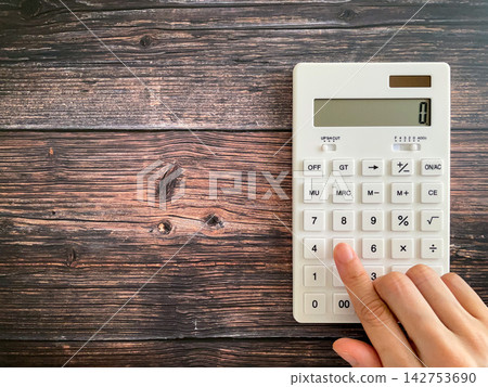 A woman's hands are calculating on a white calculator placed on a wooden table. A woman's hands are calculating on a white calculator placed on a wooden table. 142753690