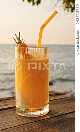 Closeup Of Tropical Drink With Straw On Wooden Table By Sea In Warm Sunlight. Summer Refreshment And Seaside Vacation Concept. Closeup Of Tropical Drink With Straw On Wooden Table By Sea In Warm Sunlight. Summer Refreshment And Seaside Vacation Concept. 142753789