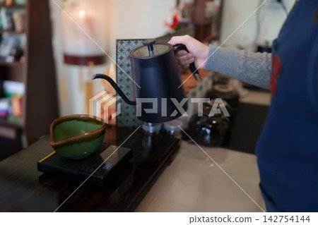 Cafe employee preparing traditional matcha pour over using gooseneck kettle into ceramic bowl at coffee shop counter Cafe employee preparing traditional matcha pour over using gooseneck kettle into ceramic bowl at coffee shop counter 142754144