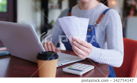 Adult cafe employee reviewing payment receipts while working on laptop with coffee at cafe table Adult cafe employee reviewing payment receipts while working on laptop with coffee at cafe table 142754147