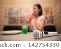 A blurred karaoke room scene with a microphone in the foreground and a woman singing in the background. 142754330