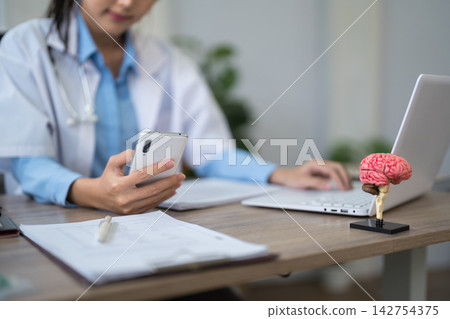 Asian female doctor reviewing patient notes on smartphone at modern clinic desk with brain model and laptop Asian female doctor reviewing patient notes on smartphone at modern clinic desk with brain model and laptop 142754375