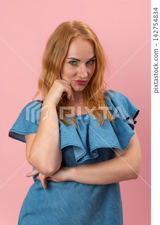 Woman with red hair and green eyes smirking playfully, holding a hand to her chin while posing with a confident and skeptical expression against a pink background Woman with red hair and green eyes smirking playfully, holding a hand to her chin while posing with a confident and skeptical expression against a pink background 142754834