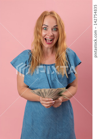 Excited woman with wide eyes and an open mouth celebrating a financial win, holding a fan of United States currency banknotes against a pink background Excited woman with wide eyes and an open mouth celebrating a financial win, holding a fan of United States currency banknotes against a pink background 142754835