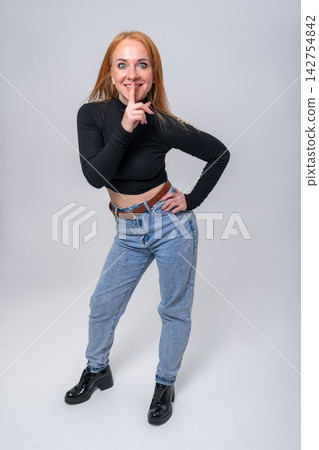 Young woman with red hair standing against a plain background, looking at the camera and making a shhh gesture, symbolizing quiet, privacy, and confidential information Young woman with red hair standing against a plain background, looking at the camera and making a shhh gesture, symbolizing quiet, privacy, and confidential information 142754842