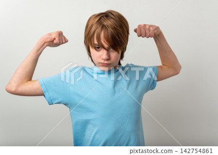 Young preteen boy in a light blue tee flexing biceps and staring confidently at the camera, conveying strength, determination and self esteem on a clean white background 142754861