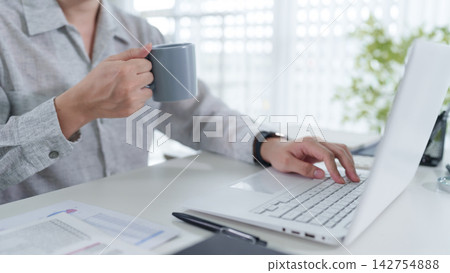 Adult woman holding coffee mug while typing on laptop at bright home office desk with copy space Adult woman holding coffee mug while typing on laptop at bright home office desk with copy space 142754888