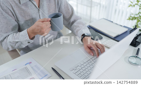 Adult professional working on laptop and holding coffee mug at bright home office desk with copy space 142754889
