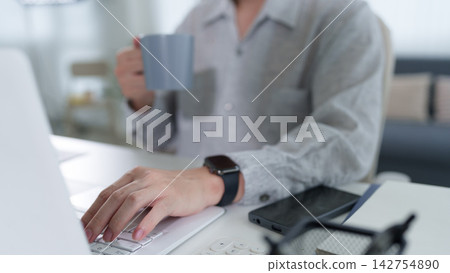 Professional woman typing on laptop while holding coffee mug at home workspace with smartphone and smartwatch visible Professional woman typing on laptop while holding coffee mug at home workspace with smartphone and smartwatch visible 142754890